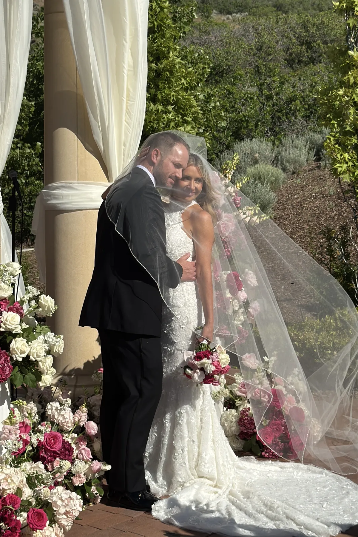 Bride and groom under a veil beside pillars and rose arrangements.