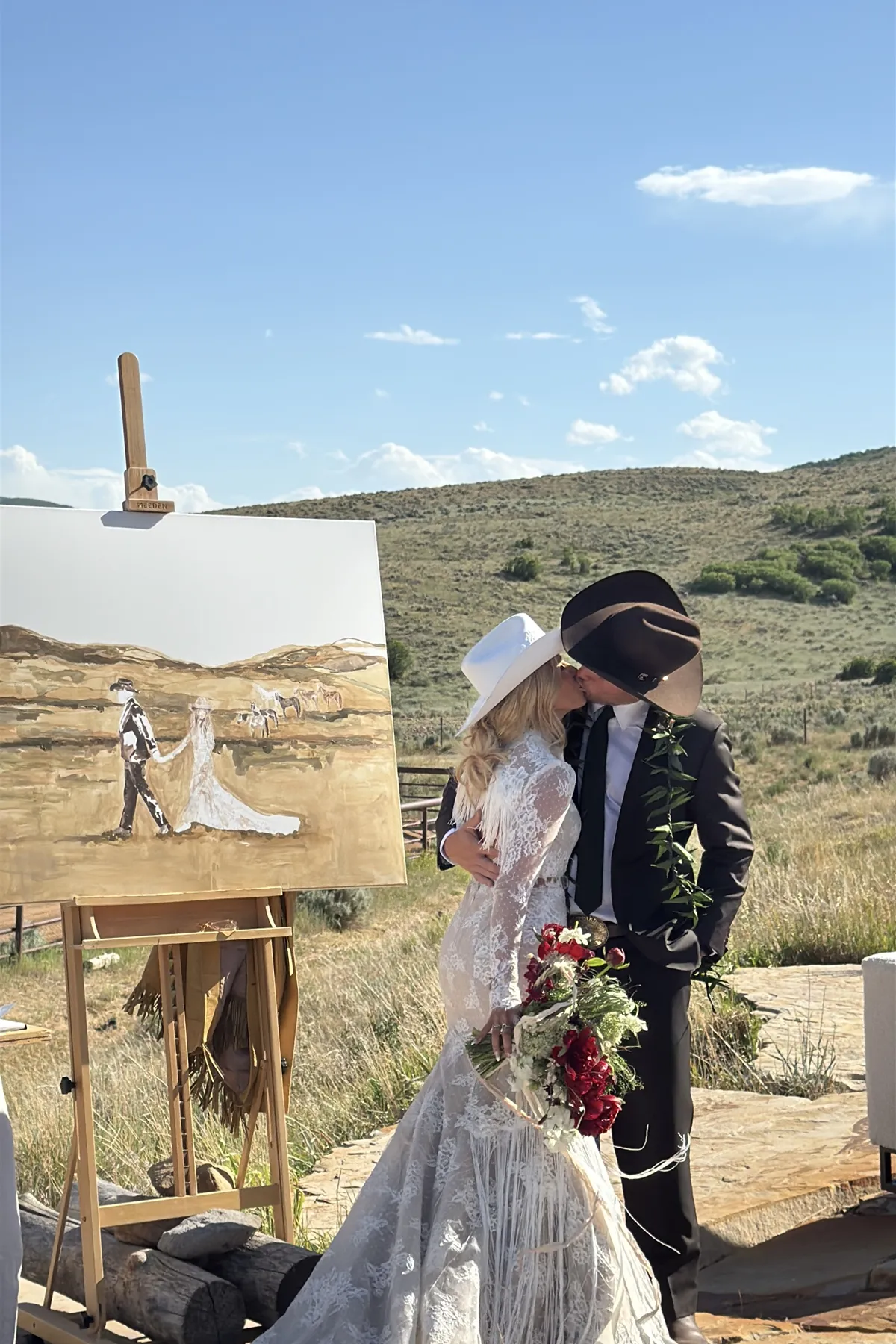 Couple kissing beside a live wedding painting at a ranch celebration.