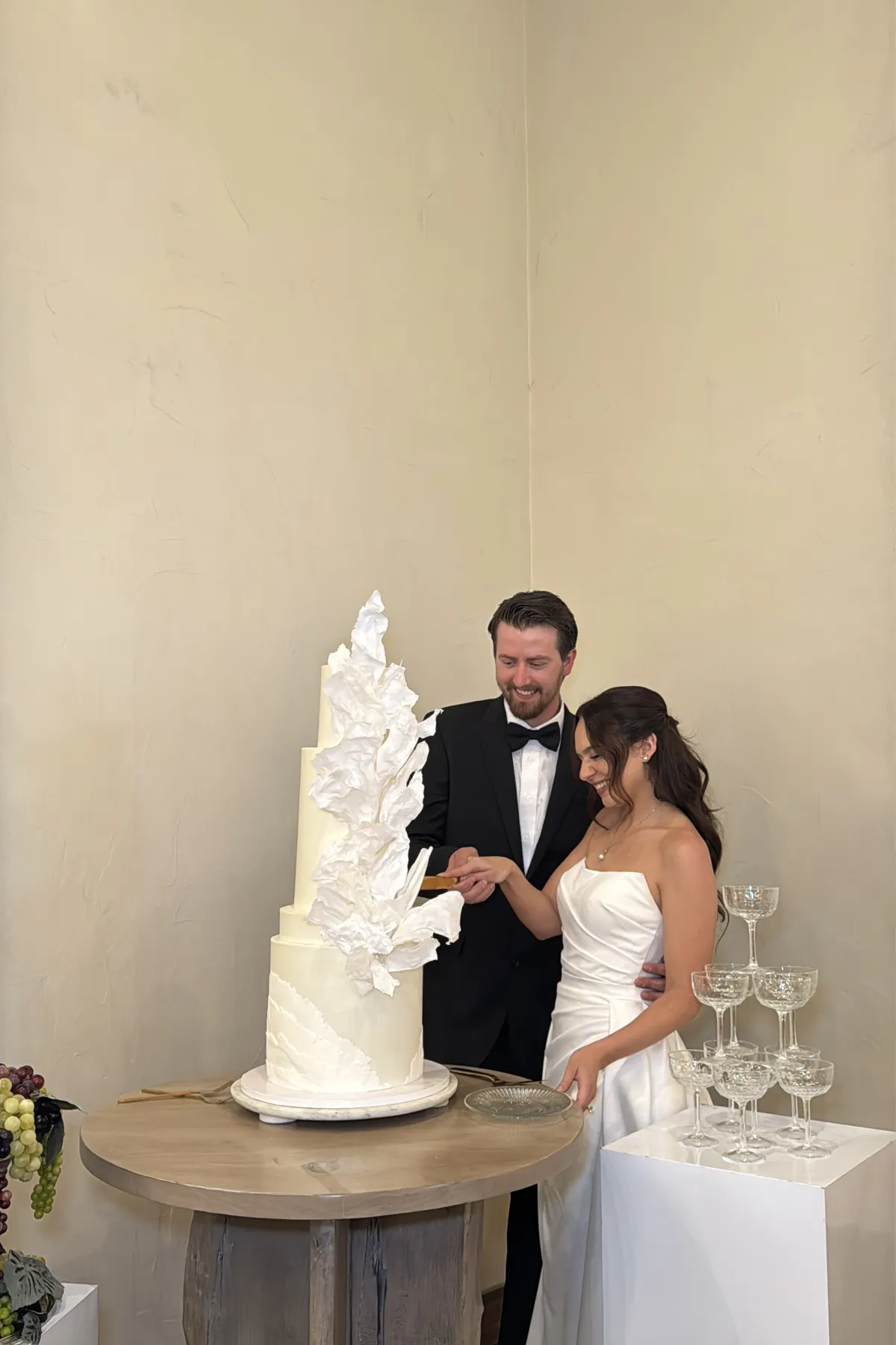 Bride and groom cutting a sculptural white cake beside a champagne tower.