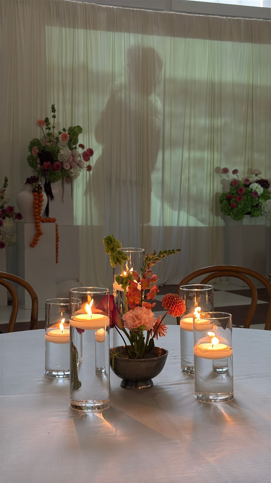 A table with floating candles and floral centerpiece, shadowed figure behind curtain