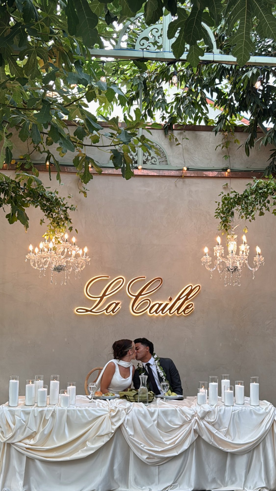 A couple kissing at a draped white head table under chandeliers