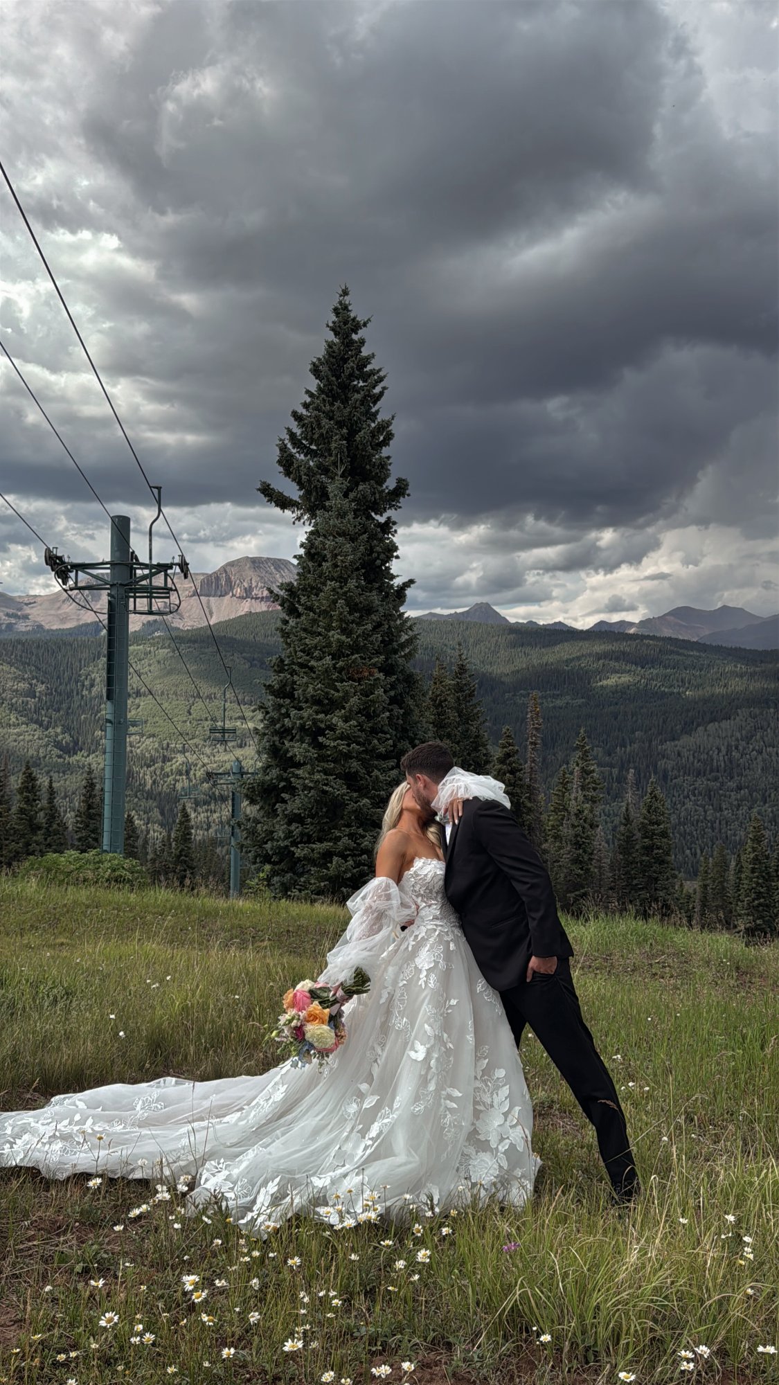 Bride and groom kissing in a mountain wildflower meadow beside a ski lift.