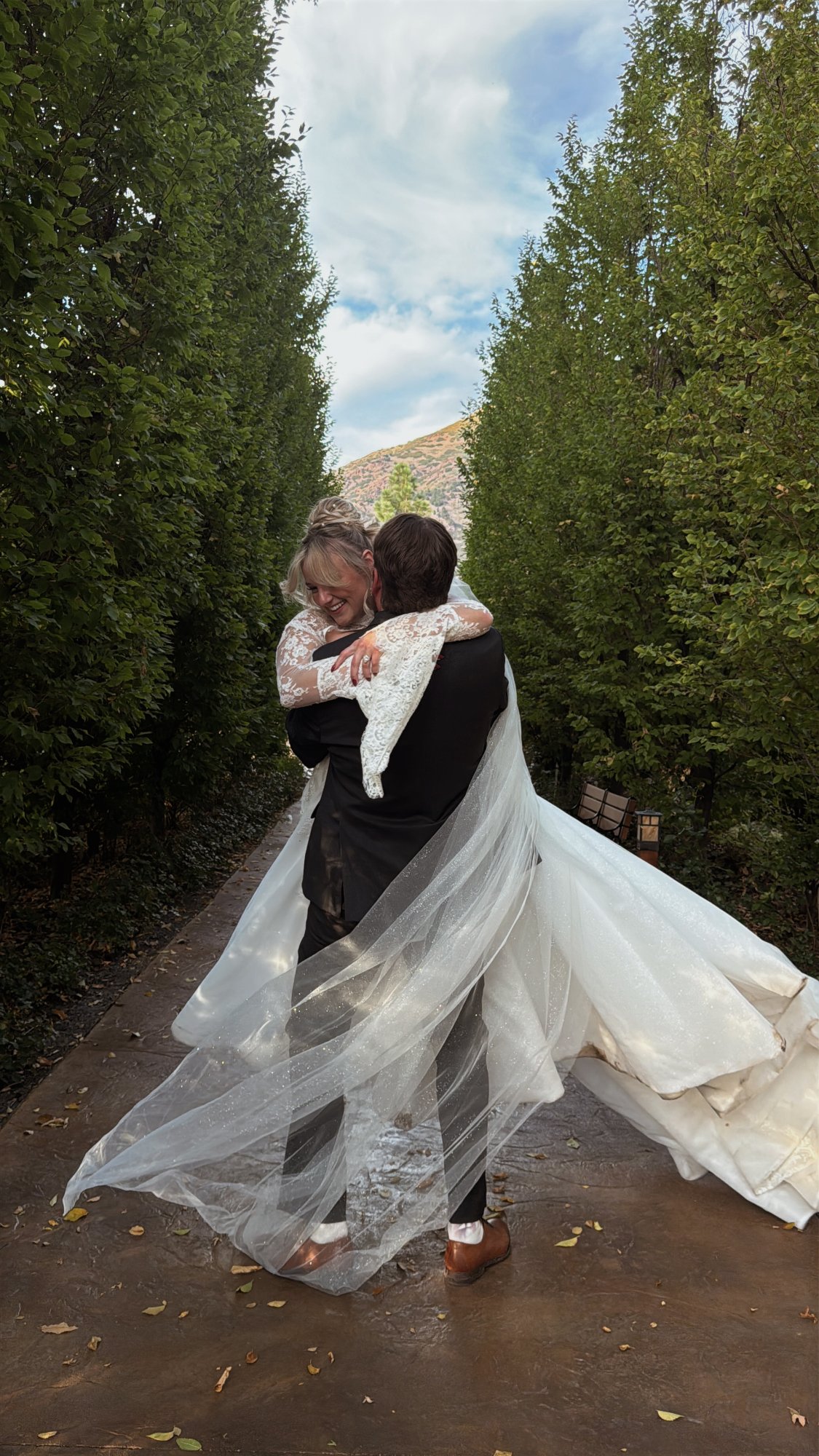 Two people embracing on tree lined path, one in white wedding gown and veil